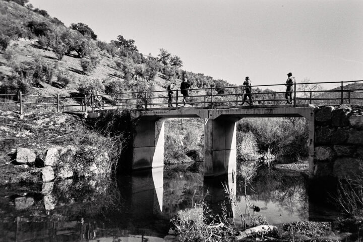 A black and white image of a concrete bridge over a calm stream, with three people walking across. The bridge is surrounded by rocky terrain and sparse vegetation, while reflections of the bridge and people can be seen in the water below.
