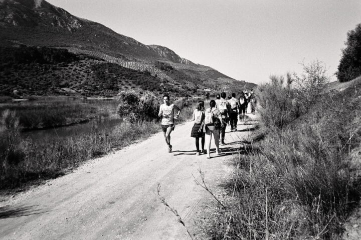 A group of runners and walkers follow a dirt path alongside a river, surrounded by scenic mountains and greenery, capturing a sense of outdoor adventure and camaraderie in a black-and-white setting.