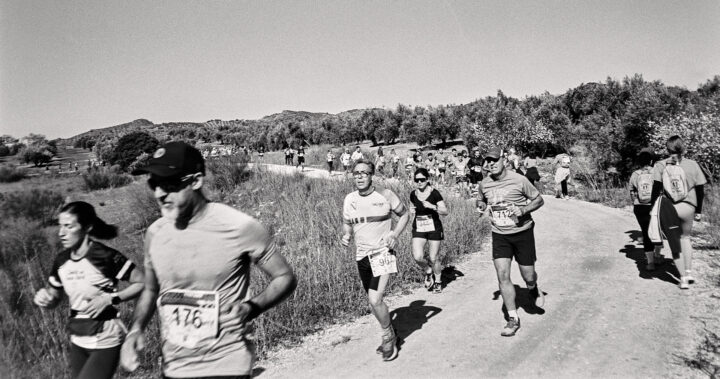 A group of runners participates in a race on a dirt path surrounded by greenery and hills, with a mix of men and women wearing numbered bibs and athletic gear under clear skies.