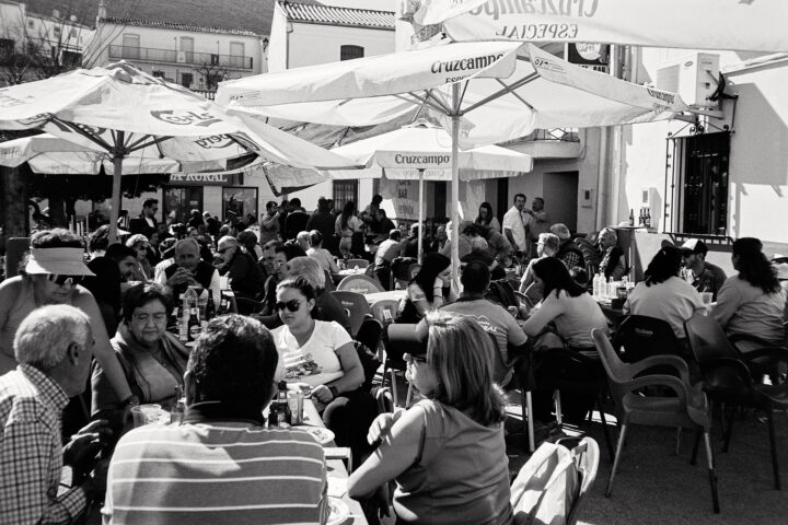 A bustling outdoor café scene in black and white, filled with people socializing under umbrellas. Tables are occupied with diverse groups enjoying food and drinks in a lively atmosphere, suggesting a vibrant community gathering.