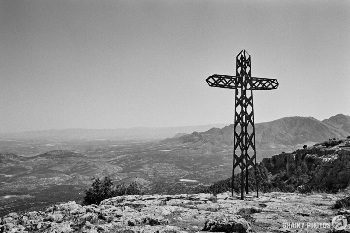 A tall metal cross, Cruz de la Atalaya, stands on a rocky hillside, overlooking a vast valley filled with mountains and greenery under a cloudy sky. The scene is captured in black and white, emphasizing the stark contrast between the cross and the landscape.