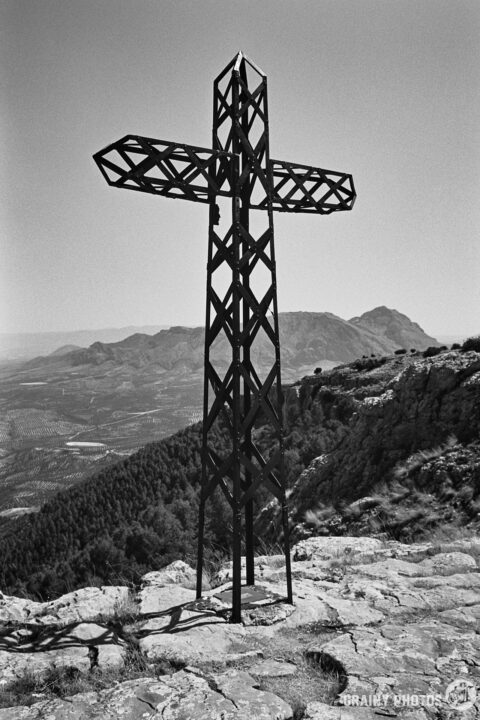 A tall, metal cross, Cruz de la Atalaya, stands on a rocky cliff, overlooking a panoramic view of rolling hills and valleys under a clear sky. The scene is captured in black and white, highlighting the contrast between the cross and the natural landscape.