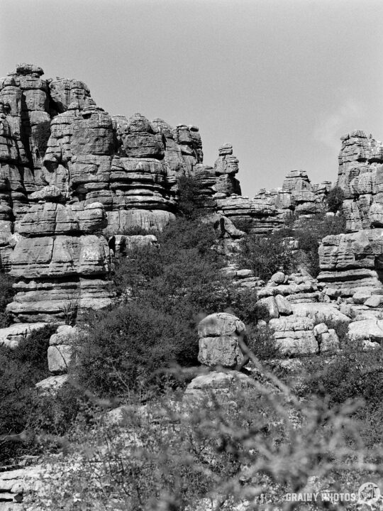 A dramatic landscape featuring towering rock formations and steep cliffs, interspersed with patches of vegetation, set against a clear sky. The scene conveys a sense of natural grandeur and rugged beauty.