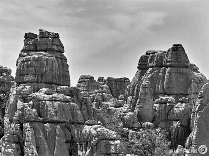 A dramatic landscape in El Torcal featuring towering karst limestone rock formations and boulders, captured in black and white. The rugged terrain showcases an intricate arrangement of stone structures against a slightly cloudy sky.