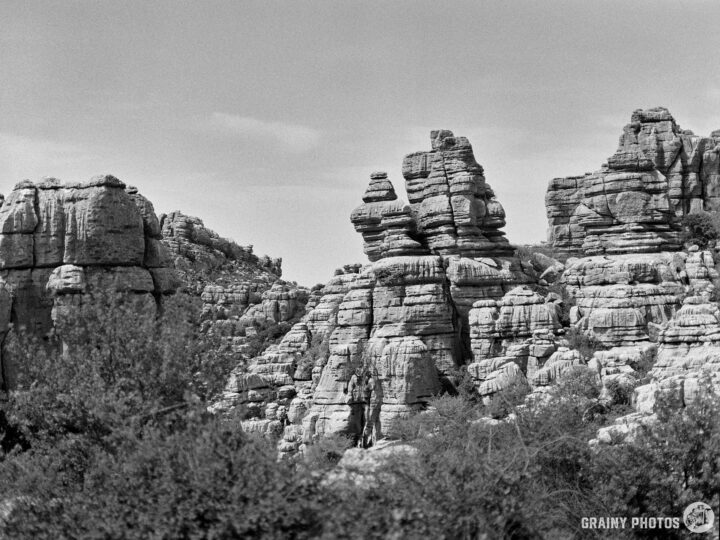 A dramatic arrangement of jagged rock formations towers against a clear, cloudy sky, surrounded by sparse vegetation. The monochrome image highlights the textures and shapes of the natural landscape.