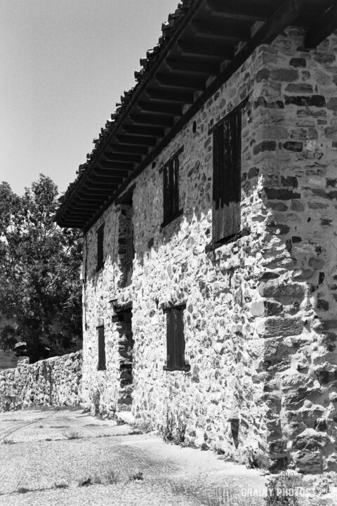 A black and white photograph of a rustic stone building with wooden accents, featuring multiple windows and surrounded by greenery, evoking a sense of historical charm and tranquility.