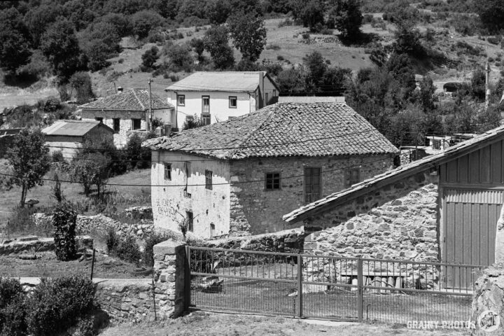 A serene black-and-white image of a rural landscape featuring traditional stone houses with tiled roofs, surrounded by greenery and rolling hills, offering a glimpse into rustic life.