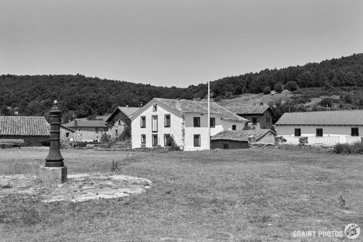 The village green in a rural landscape featuring a small village with traditional houses, a flagpole, and a stone well in a grassy area, surrounded by hills and trees.