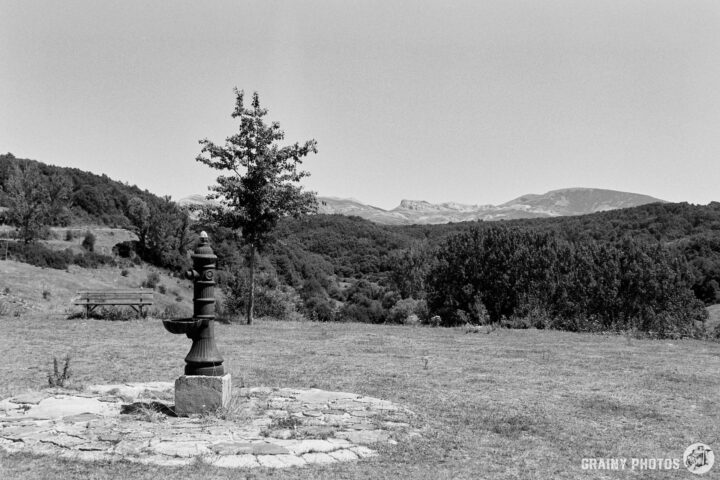 A vintage water pump stands on a stony base in a serene countryside setting, surrounded by lush greenery and rolling hills under a clear sky. A picnic table is visible in the distance, adding to the tranquil rural atmosphere.