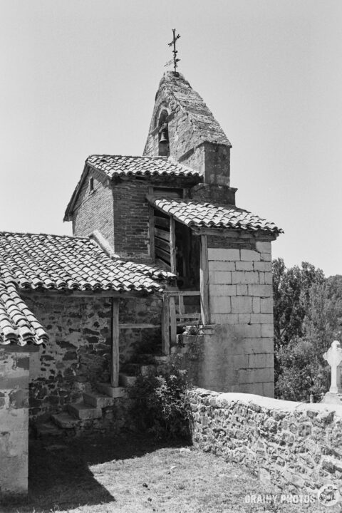 A black and white photograph of a rustic stone church, Nuestra Señora de la Asunción, with a bell tower topped by a cross. The building features a tiled roof and is surrounded by greenery, with a stone staircase leading to the entrance.