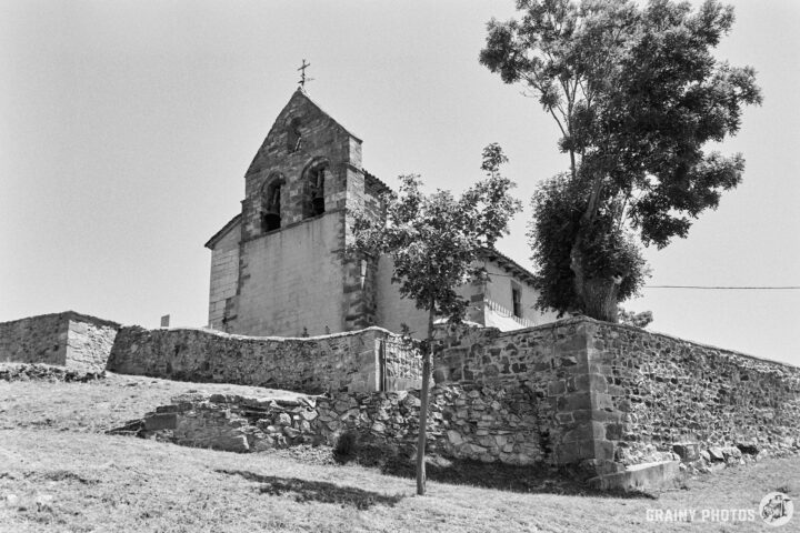 The quaint Iglesia de Nuestra Señora de la Asunción church stands atop a grassy hill, surrounded by stone walls and trees, with a clear sky in the background. The structure features a triangular roof and a cross, showcasing rustic architecture in a serene landscape.