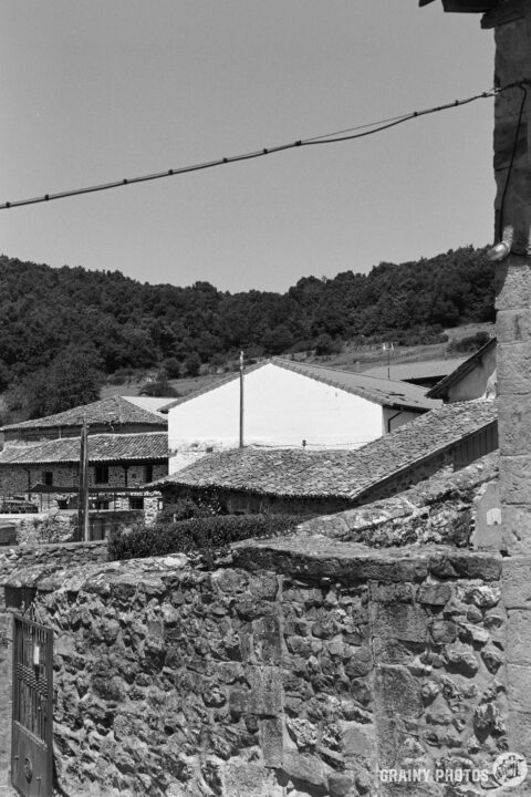 A scenic black-and-white view of traditional stone houses, with sloped roofs and greenery in the background, evoking a rustic village atmosphere. The image captures the charm of rural architecture against a clear sky.