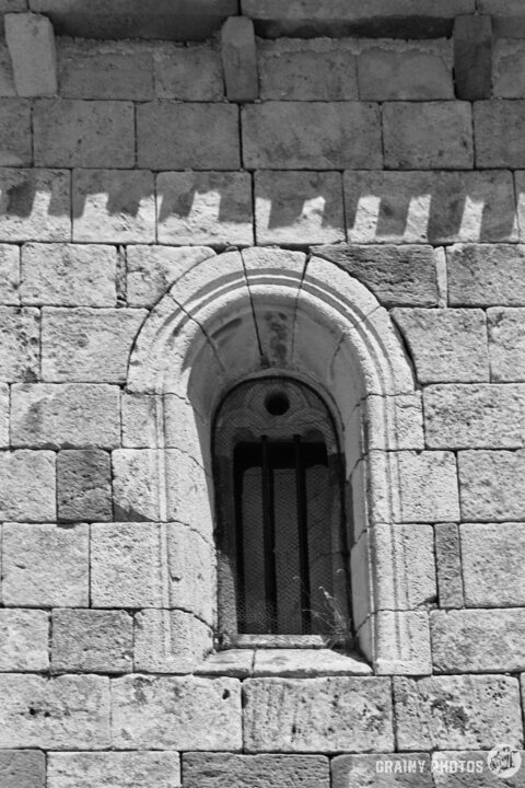 A black and white image of a stone wall in Iglesia de Nuestra Señora de la Asunción church. featuring an arched window with metal bars. The textured stones are well-defined, showcasing architectural details typical of a historic church.
