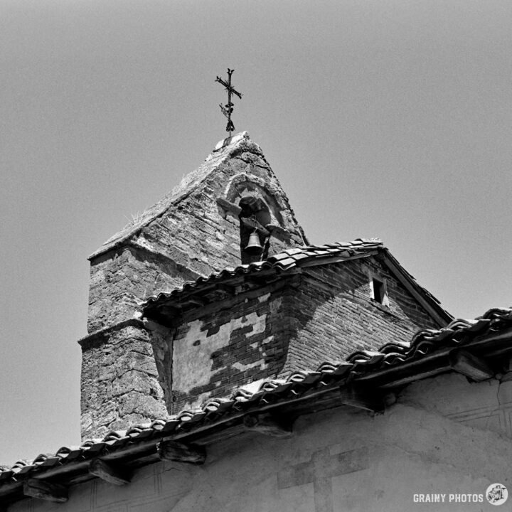 A black and white photo of the Iglesia de Nuestra Señora de la Asunción church steeple in Estalaya featuring a cross at the top, with an intricately designed roof and textured stone walls, highlighting architectural details against a clear sky.