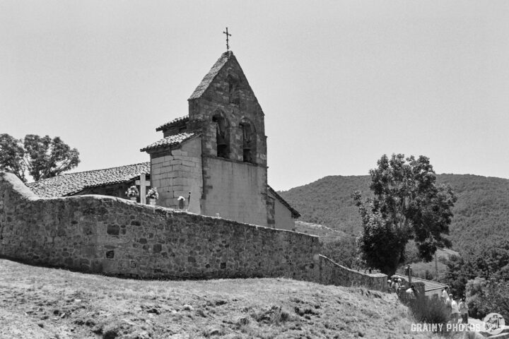 Iglesia de Nuestra Señora de la Asunción church with a bell tower, nestled on a hillside, surrounded by trees and mountains in a black-and-white photograph. The scene captures the serene landscape and architectural charm of this ancient structure.