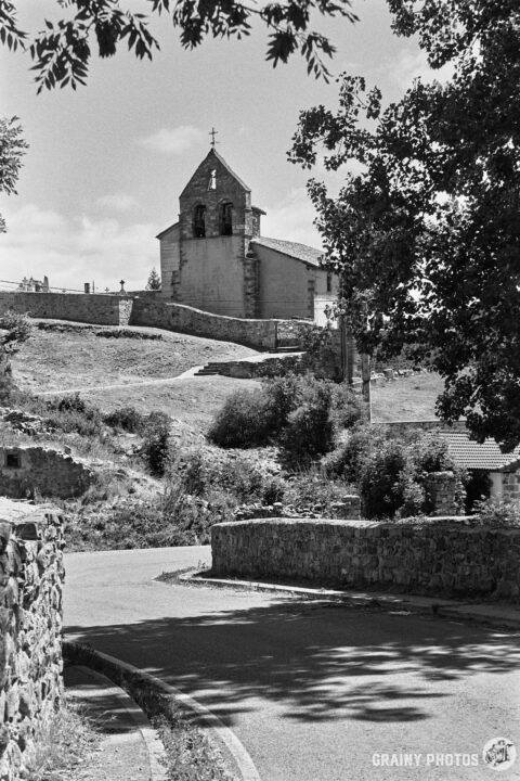 A winding road leads to the Estalaya village church situated on a hill, surrounded by lush greenery and stone walls. The scene captures a serene countryside landscape in black and white.