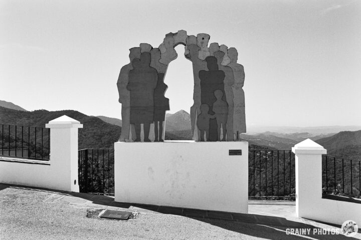 A black and white image of a sculptural monument featuring abstract figures, standing on a terrace overlooking rolling hills and a distant landscape. The scene captures a serene view, framed by a white railing and stone pathway.
