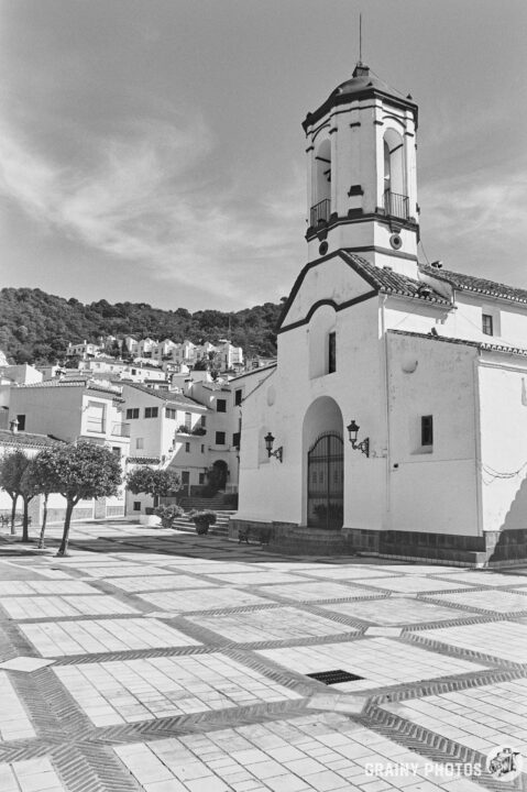 A black-and-white photo of a quaint village square featuring St. Peter the Martyr of Verona church with a prominent bell tower, surrounded by trees and traditional architecture, set against a hilly backdrop under a partly cloudy sky.