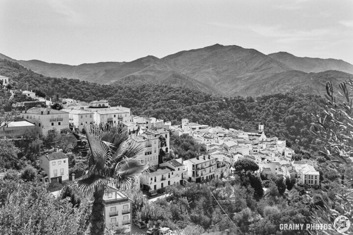 A scenic black and white view of a mountainous landscape, showcasing Genalguacil village with white buildings nestled among lush greenery, framed by towering mountains in the background.