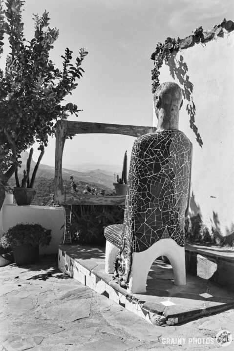 A black-and-white photo of a mosaic chair sculpture facing a framed view. Surrounding it are potted plants and a backdrop of mountains, creating a serene artistic atmosphere.