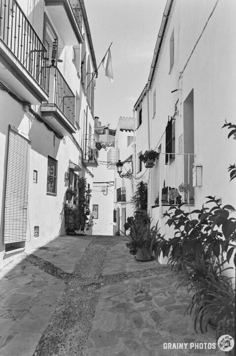 A narrow, winding alleyway lined with whitewashed buildings and blooming plants. Balconies adorned with pots and a flag fluttering above, creating a charming, quaint atmosphere typical of Mediterranean architecture.