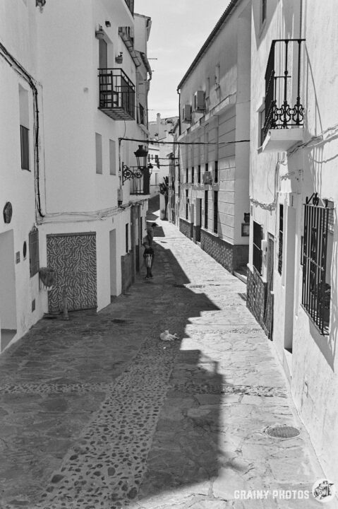 A narrow cobblestone alleyway lined with whitewashed houses featuring balconies. A solitary figure walks down the street under bright sunlight, creating a peaceful, picturesque scene. Shadows contrast with the sunlit stones, enhancing the atmosphere.