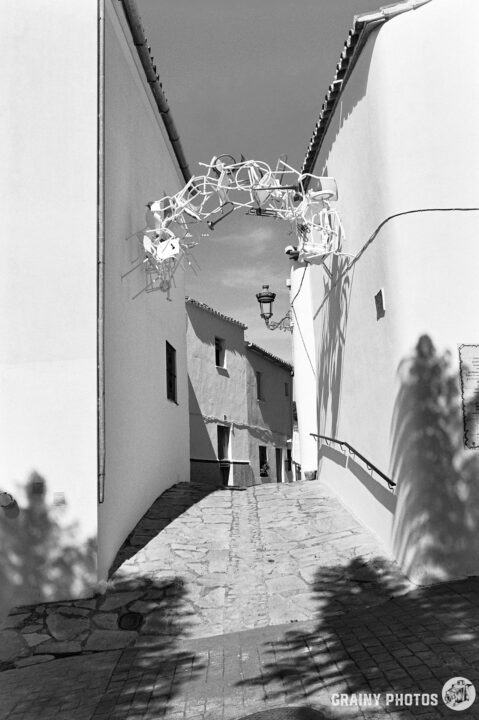 A narrow, winding street with whitewashed walls leads into a quaint alleyway. An artistic arch made of intertwined chairs hangs overhead, casting shadows on the cobblestone path. The scene is captured in black and white, enhancing its charm.