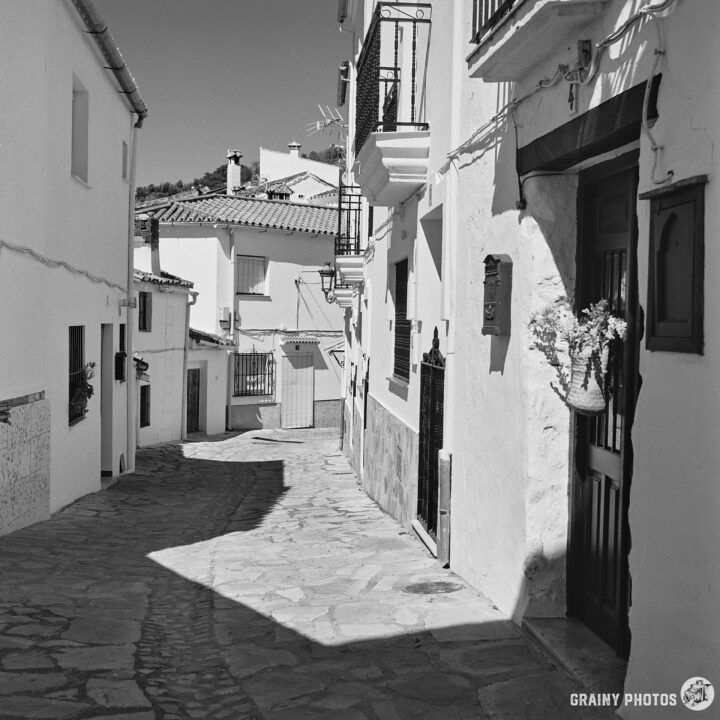A narrow cobblestone street lined with whitewashed buildings and balconies, illuminated by sunlight and casting long shadows. The scene evokes a quaint, serene atmosphere typical of Genalguacil.