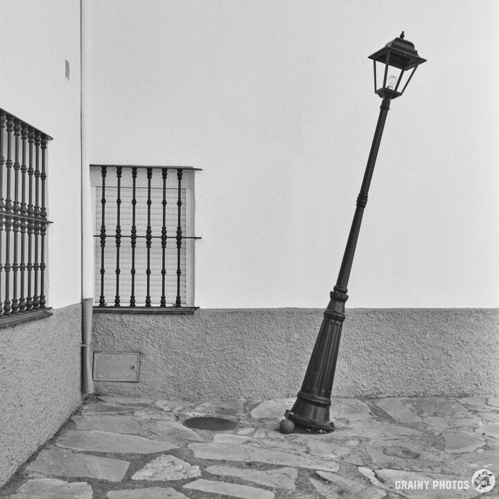 A slanted street lamp, propped up by an orange wedged under the base, stands beside a white wall, casting a unique shadow. Nearby, simple metal bars on windows add contrast to the scene. The cobblestone path enhances the rustic charm of this urban corner.