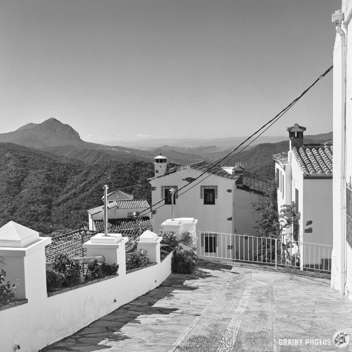 A scenic black and white view of a path leading downhill, surrounded by traditional white buildings. In the distance, rolling hills and mountains create a serene landscape under a clear sky.