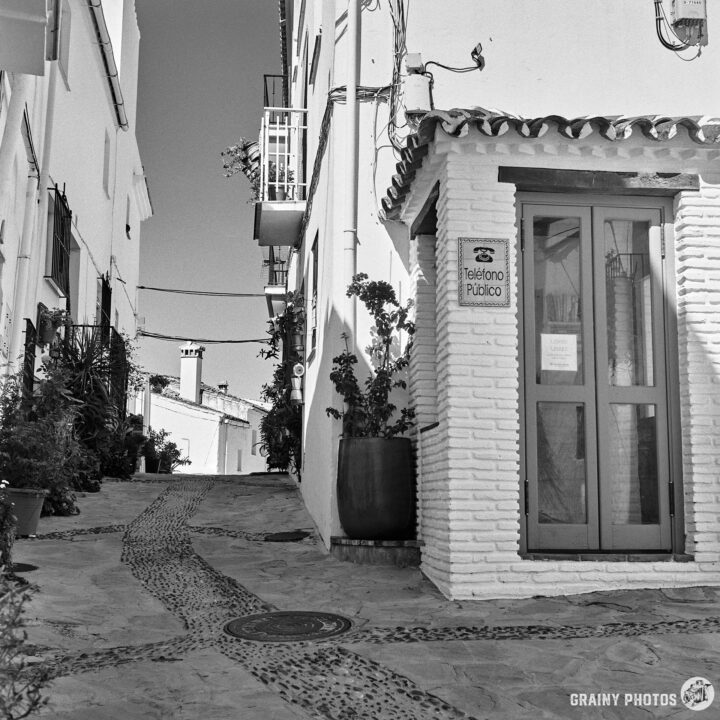 A narrow cobblestone alleyway lined with whitewashed buildings, featuring a public telephone booth (used as a mini library). Potted plants adorn the path, creating a serene, picturesque atmosphere in a quaint village setting.