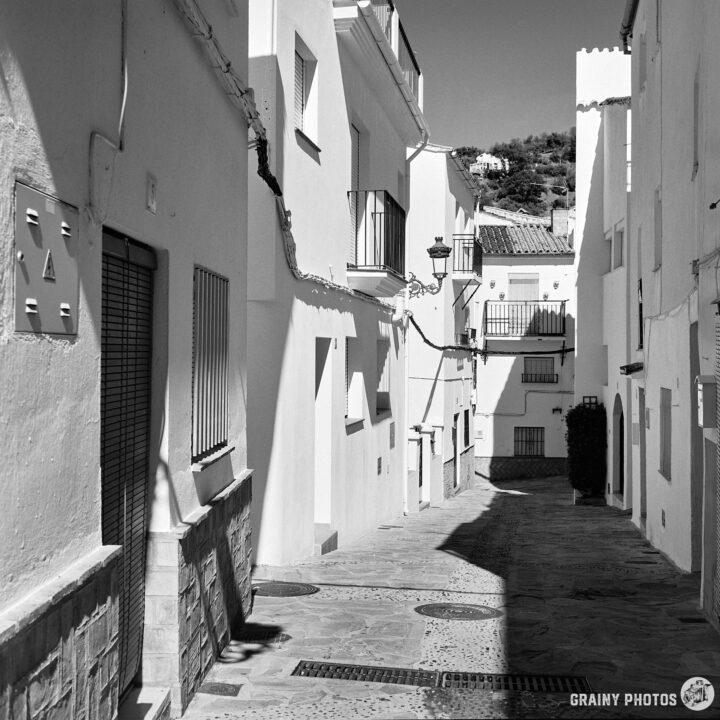 A narrow, sunlit alleyway lined with whitewashed buildings, featuring stone pavements and traditional balconies. The scene is captured in black and white, enhancing the shadows and textures of the architecture.