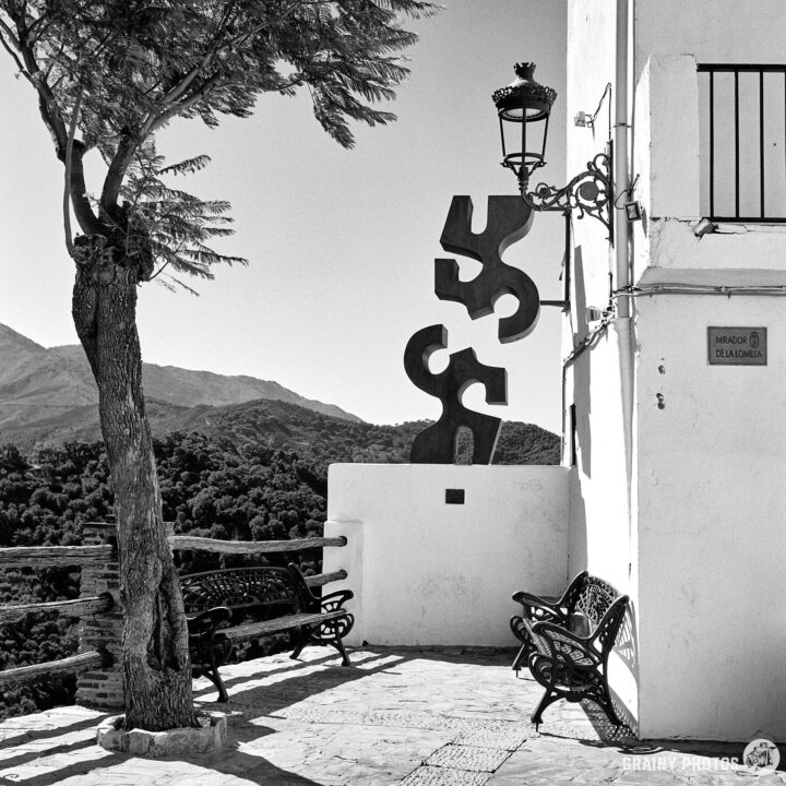 A tranquil outdoor scene featuring a black and white view of a village plaza with benches, a decorative sculpture, and a nearby tree. The backdrop includes rolling hills and a clear sky, evoking a serene atmosphere.