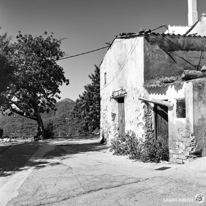 A black-and-white photograph of an abandoned stone house on the edge of Genalguacil with a wooden door, surrounded by trees and mountains. A serene, rural setting.