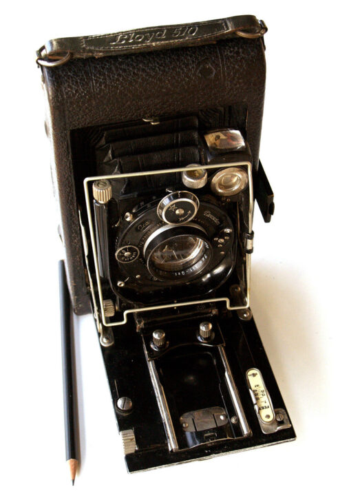 A vintage ICA Lloyd 510 camera with a folding design, featuring a large lens and metal details. It's set against a white background alongside a pencil, showcasing its classic aesthetic and functionality.