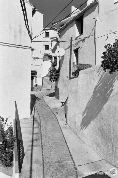 A black-and-white photo of a narrow cobbled meandering passageway in Jubrique with white hoses on both sides.