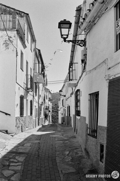 A black-and-white photo of a narrow cobbled street in Jubrique with white houses on both sides. An old-style street lantern is fixed to the first house on the right.