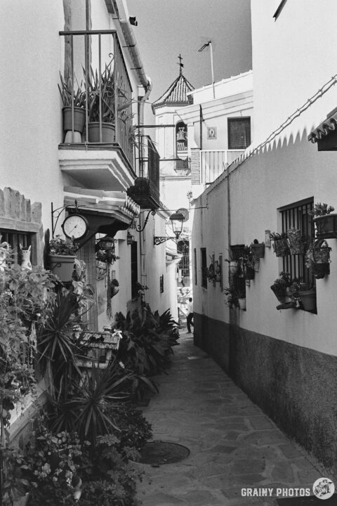 A black-and-white photo of a pretty narrow cobbled passage between white houses in Jubrique. On the left side, there are numerous large potted plants, making the passage even narrower. The town church is visible at the end of the street.