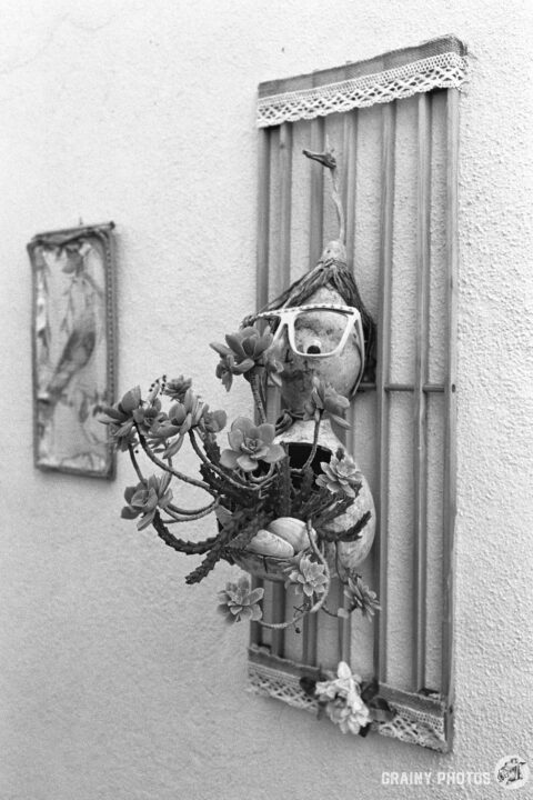 A black-and-white photo of a small pottery human caricature decorated with seashells and flowers and wearing oversized glasses. A beady eye appears to look straight at you.