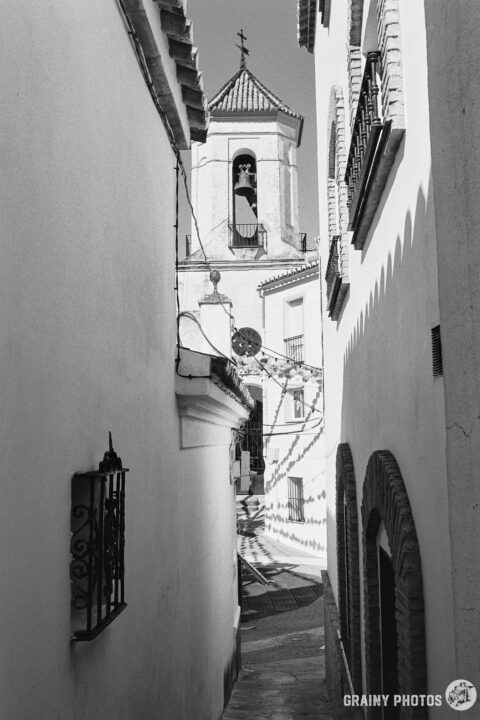 A black-and-white photo of a narrow alley with white houses on both sides leading to the town square. The church's bell tower is visible at the end of the alley.