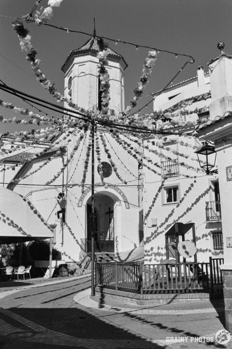 A black-and-white photo of the town square with the church in the background. There is bunting strung everywhere - it's fiesta week.