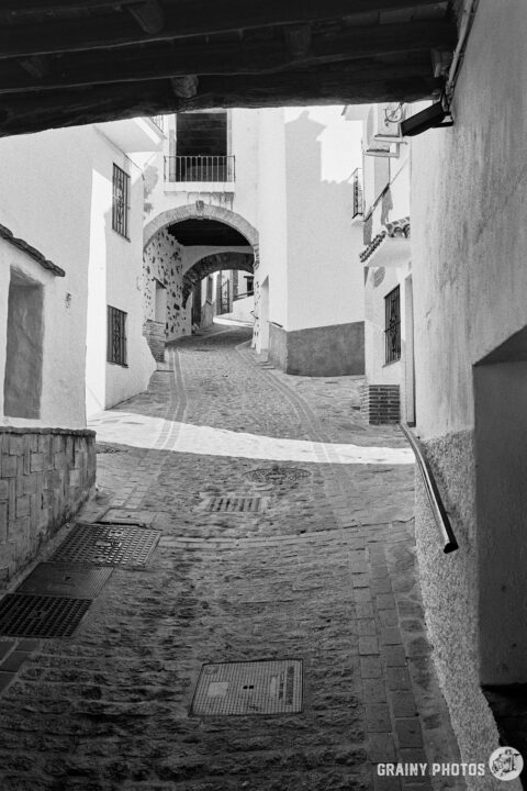 A black-and-white photo looking along a narrow, meandering, cobbled street from under a tinao.