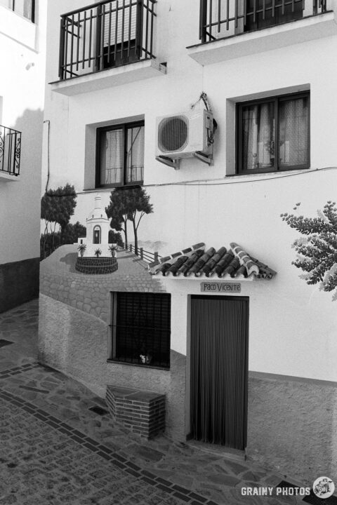 A black-and-white photo of a white town-house with a mural of a village square with a central fuente feature.