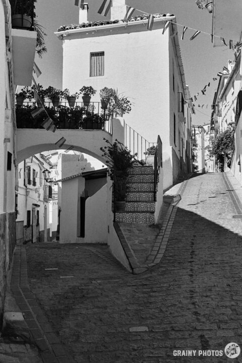 A black-and-white photo of a Y junction of a narrow cobbled street in Jubrique. The left fork heads downhill under an arched terrace.