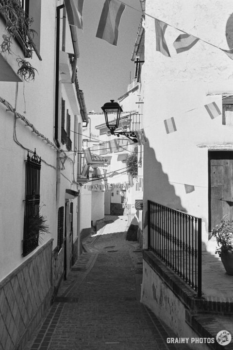 A black-and-white photo of a pretty narrow cobbled street with white houses and bunting overhead - it's fiesta week.