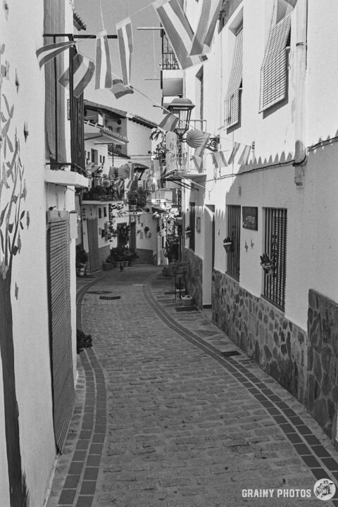 A black-and-white photo of a pretty narrow cobbled street with white houses and bunting overhead - it's fiesta week.