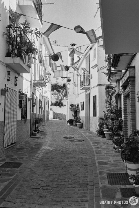 A black-and-white photo of a pretty narrow cobbled street with white houses and bunting overhead - it's fiesta week.
