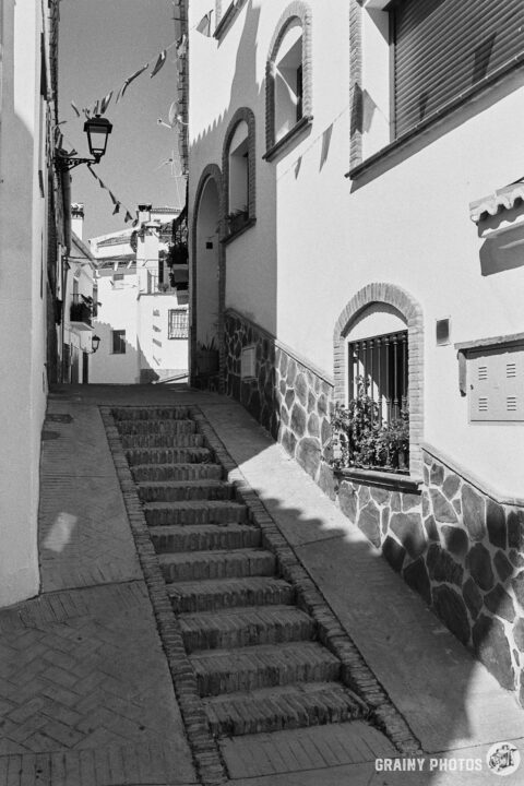 A black-and-white photo of of steps in a narrow passage between white houses. Fiesta bunting overhead.
