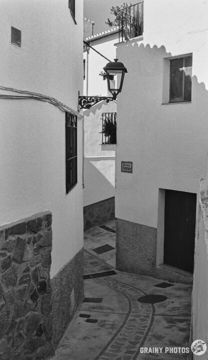 A black-and-white photo looking along a narrow alley which twists and turns between white houses.
