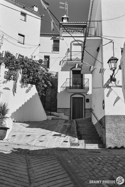 A black-and-white photo of several junctions between two parallel narrow streets on a hillside. Cobblestones, potted plants, steps, white houses and fiesta bunting - it's all in this photo!